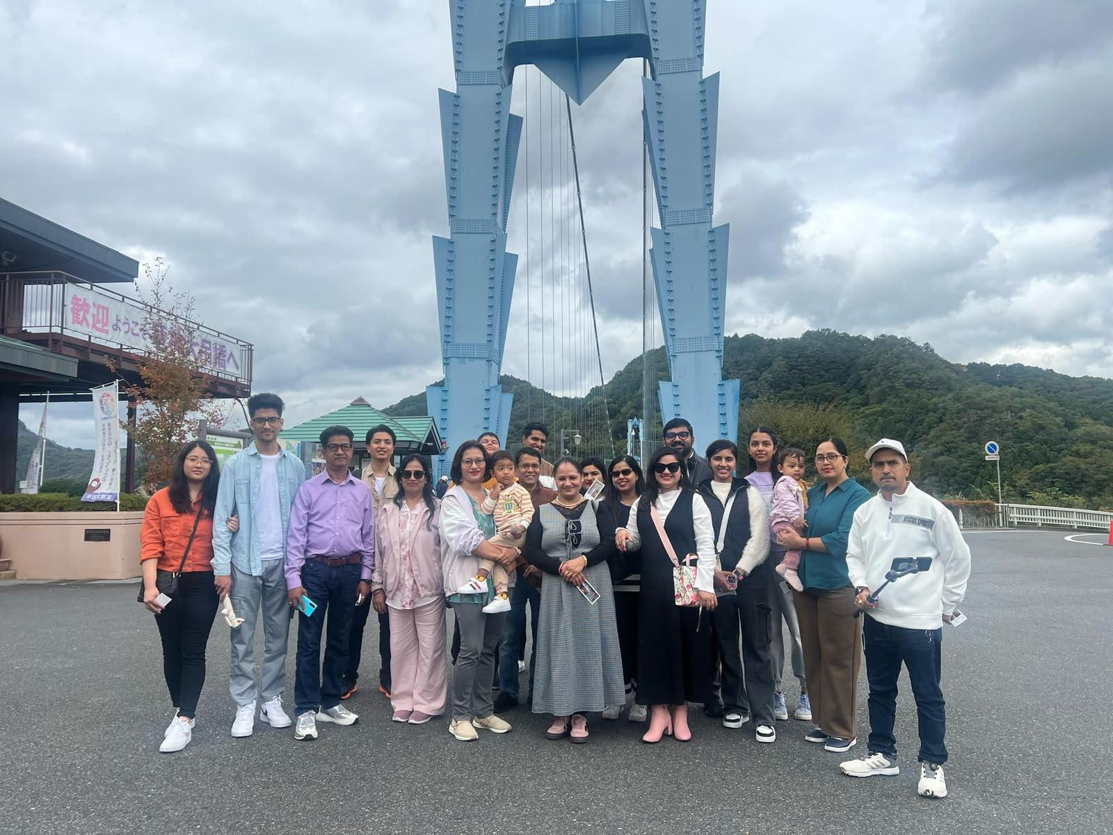 A large multi-generational Nepali group in autumn clothing in front of a blue suspension bridge